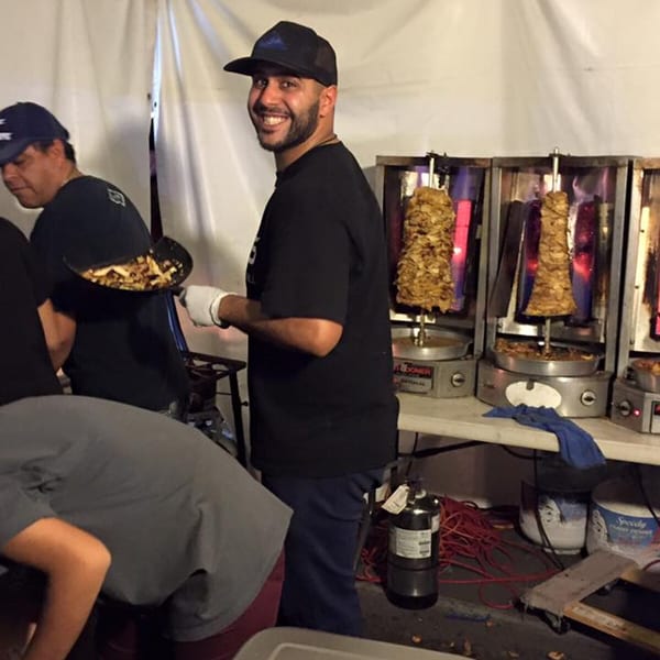 A smiling chef stands next to vertical rotisseries with Mediterranean shawarma meat cooking.