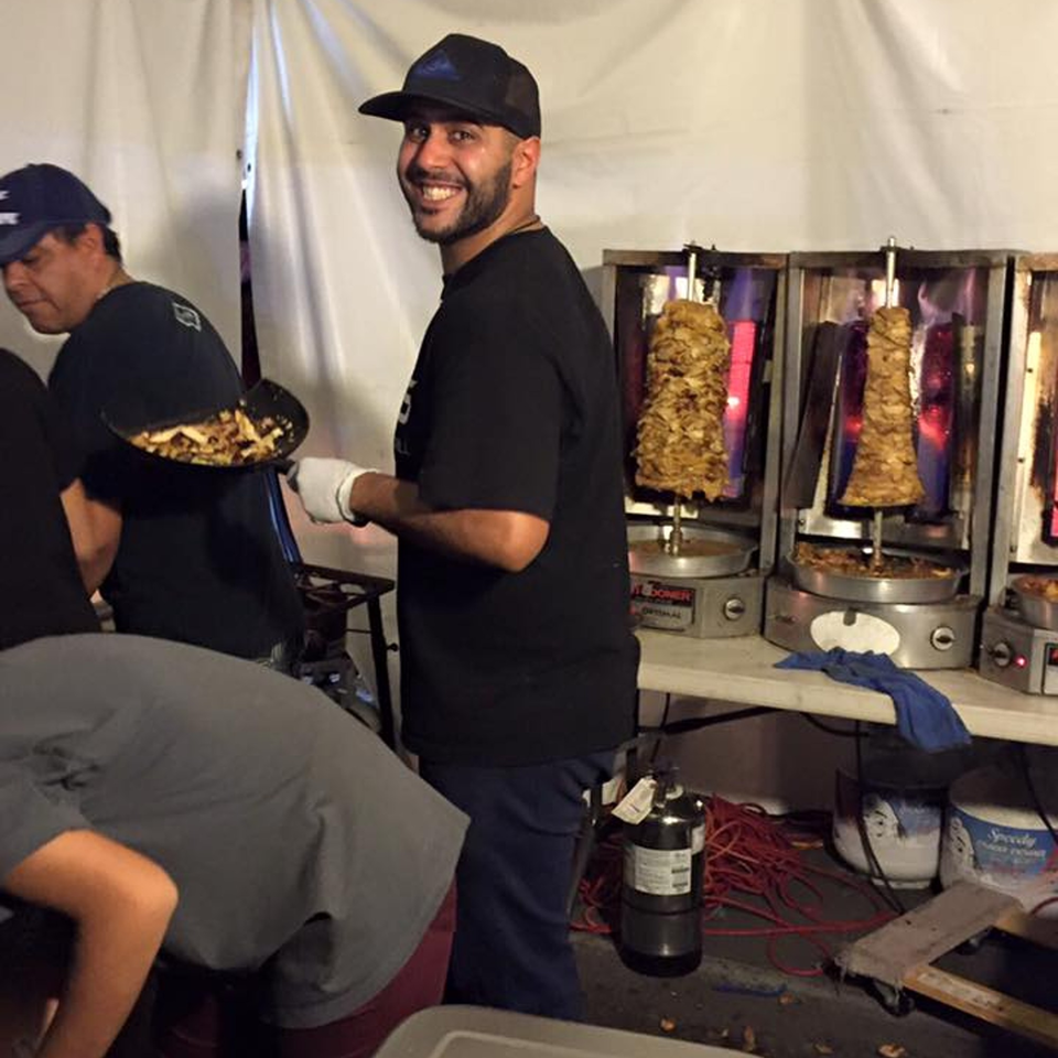A smiling chef stands next to vertical rotisseries with Mediterranean shawarma meat cooking.