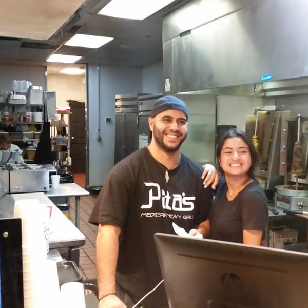 Two smiling Pita's Mediterranean Grill employees stand in a busy commercial kitchen with shawarma machines.