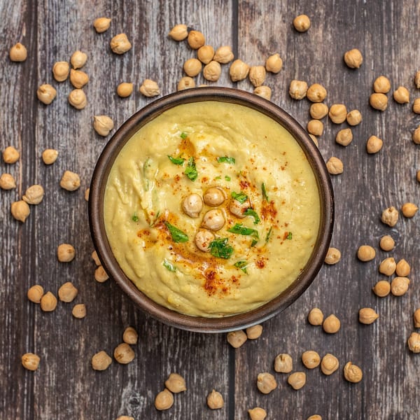 Creamy hummus in a dark bowl, garnished with chickpeas, paprika, and parsley, on a wooden surface.
