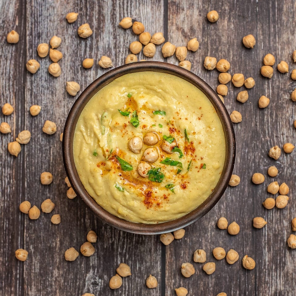 Creamy hummus in a dark bowl, garnished with chickpeas, paprika, and parsley, on a wooden surface.
