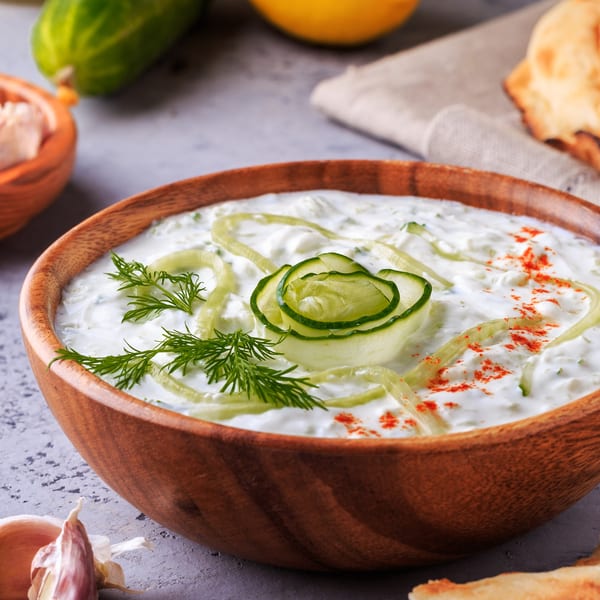 Close-up of a wooden bowl filled with creamy tzatziki, garnished with cucumber ribbons, dill, and paprika.