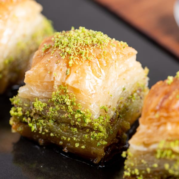 Close-up of three pieces of baklava, topped with crushed pistachios, on a dark surface.